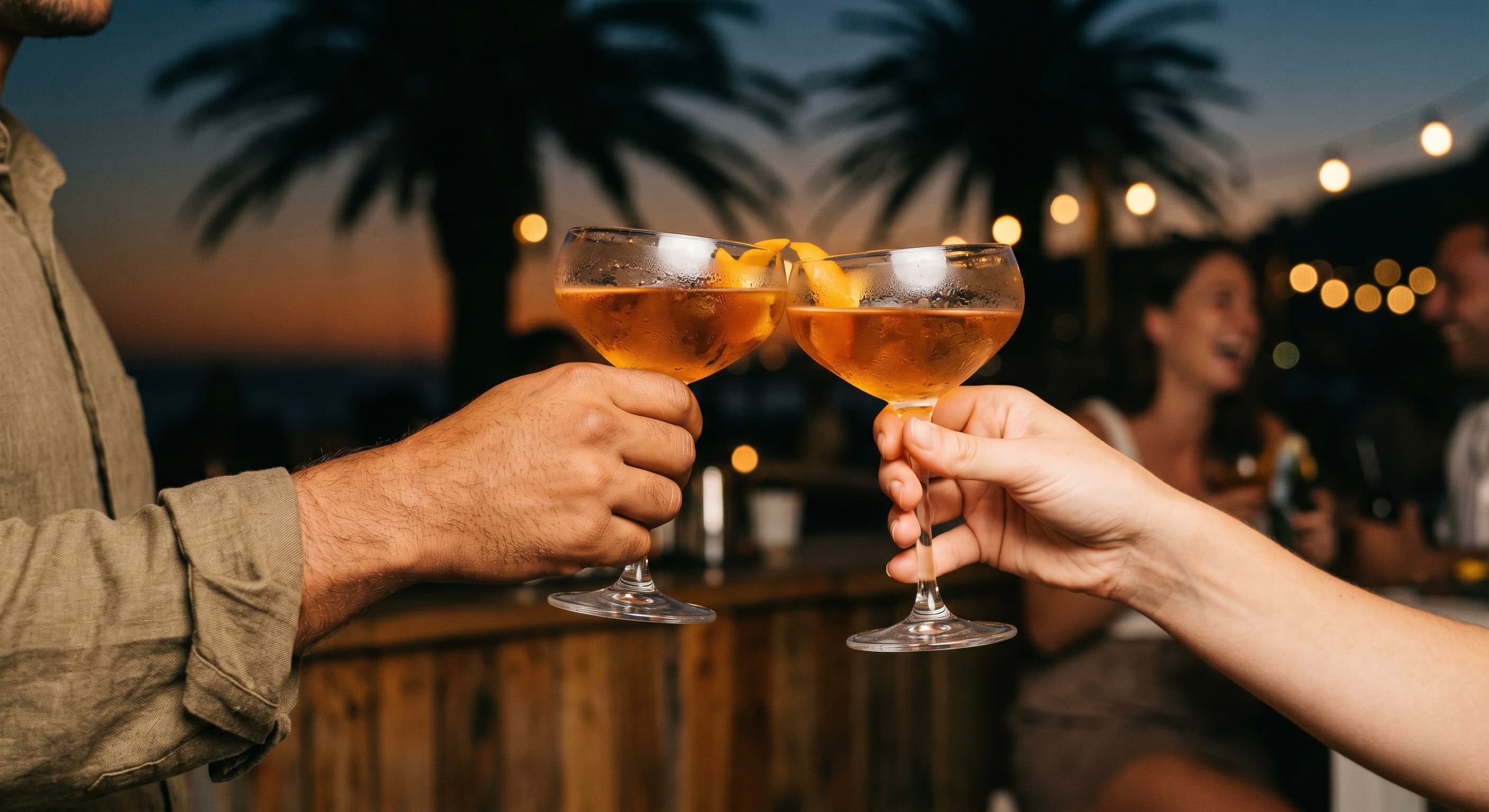 Two hands raising amber cocktail glasses in a toast, palm silhouettes and warm bokeh light flares behind them.